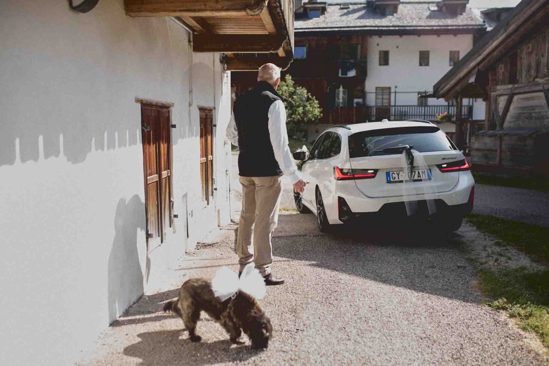 Un uomo osserva un cagnolino con fiocco bianco vicino a un'auto bianca addobbata per un matrimonio a Cortina d'Ampezzo.