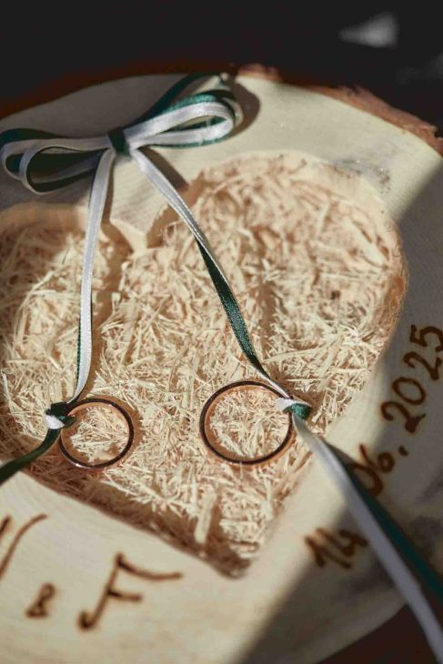 Dettaglio di due fedi nuziali in oro poggiate su un portafedi in legno a forma di cuore con incisa la data 14.06.2025.