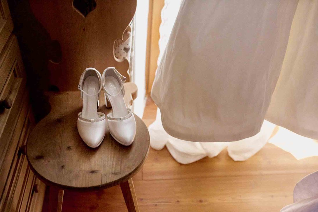 Scarpe da sposa su sedia in legno e abito bianco, dettaglio fotografato da un fotografo matrimonio Cortina d’Ampezzo.