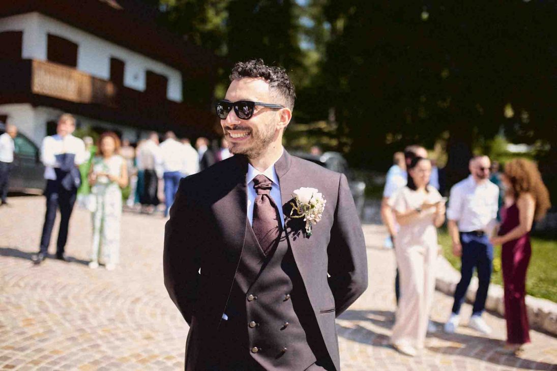 Lo sposo in abito scuro con occhiali da sole e fiore all'occhiello sorride all'aperto durante il ricevimento a Cortina.
