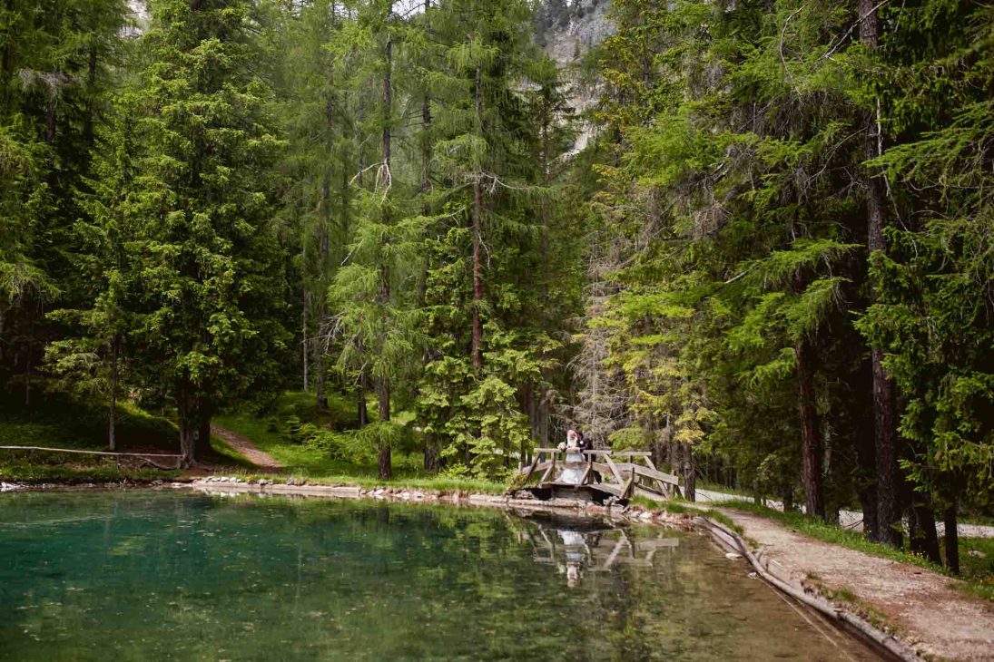 Sposi su un ponte di legno in un bosco alpino, ritratti da un fotografo matrimonio Cortina d’Ampezzo presso un lago.