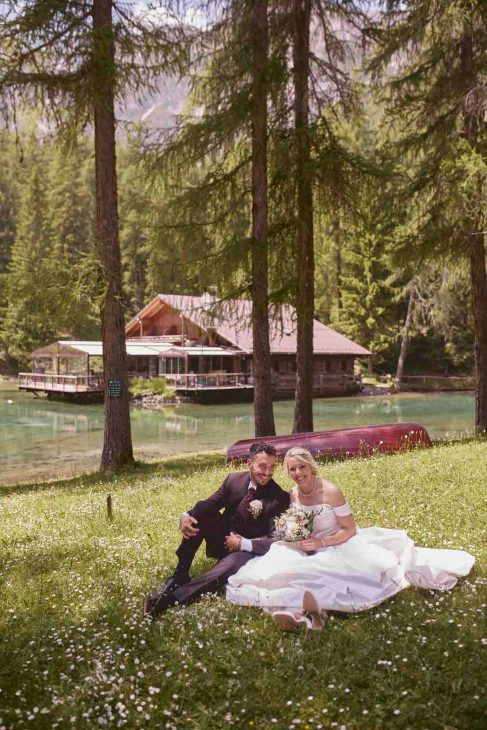 Sposi seduti su un prato fiorito davanti a uno chalet sul lago, scatto di un fotografo matrimonio Cortina d’Ampezzo.