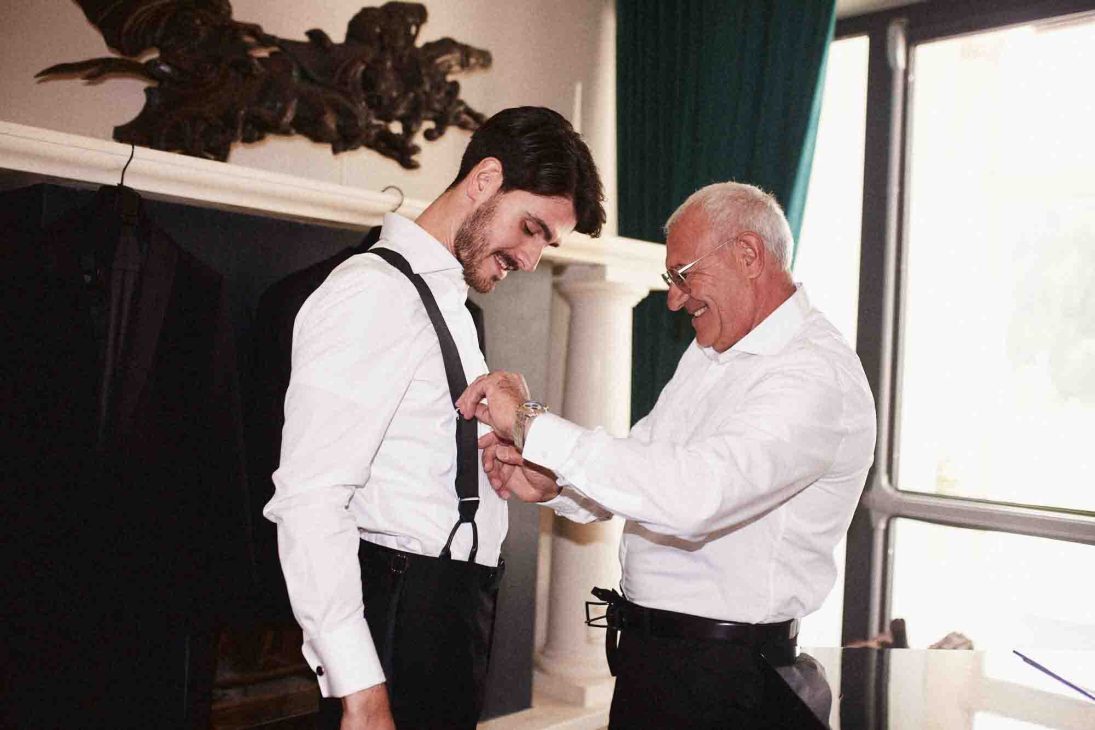 Fotografo matrimonio villa veneta Vicenza: lo sposo aiutato con le bretelle durante la preparazione in una sala storica.